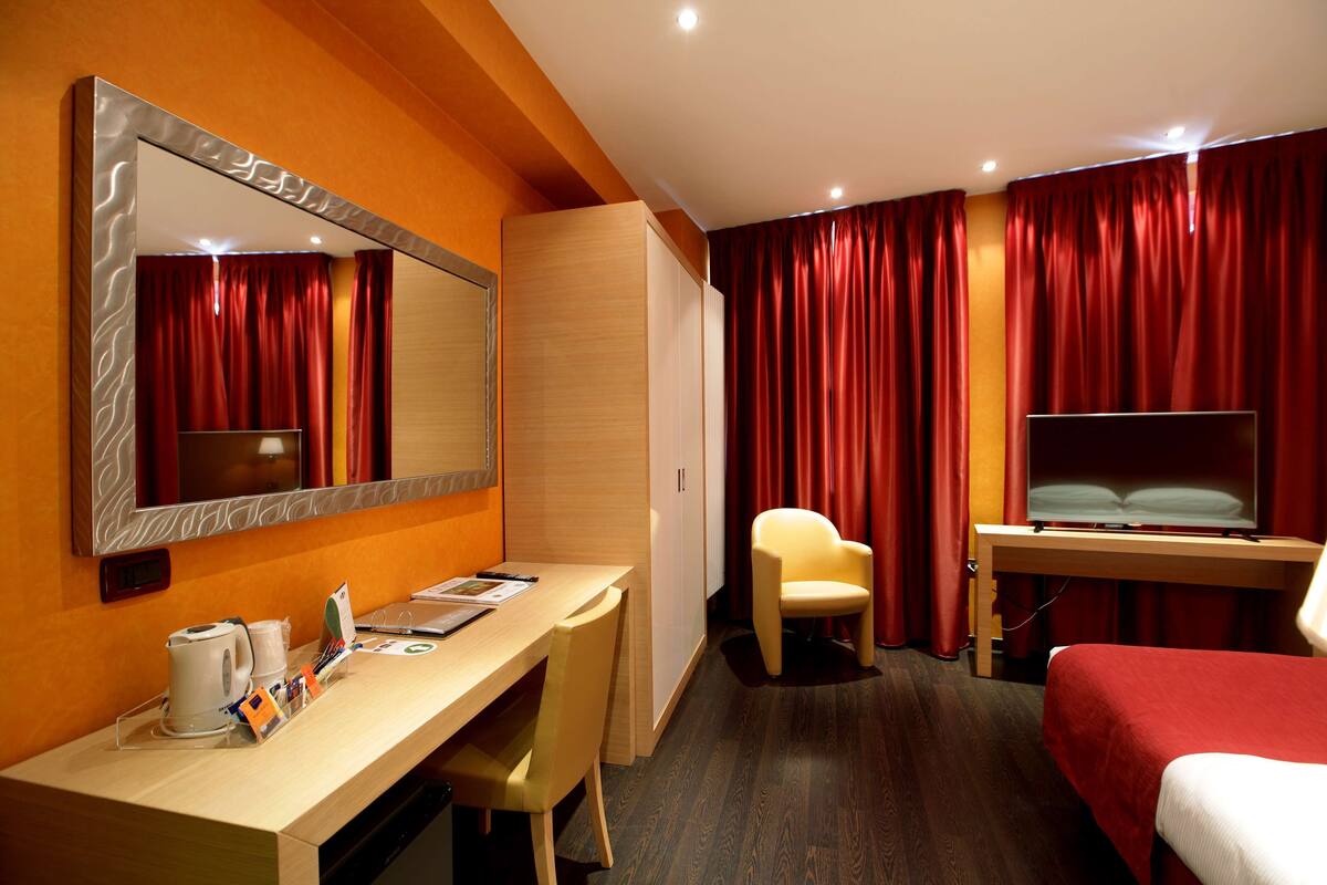 Una habitación de hotel con un escritorio de madera, una silla, una cama, un televisor y cortinas rojas.