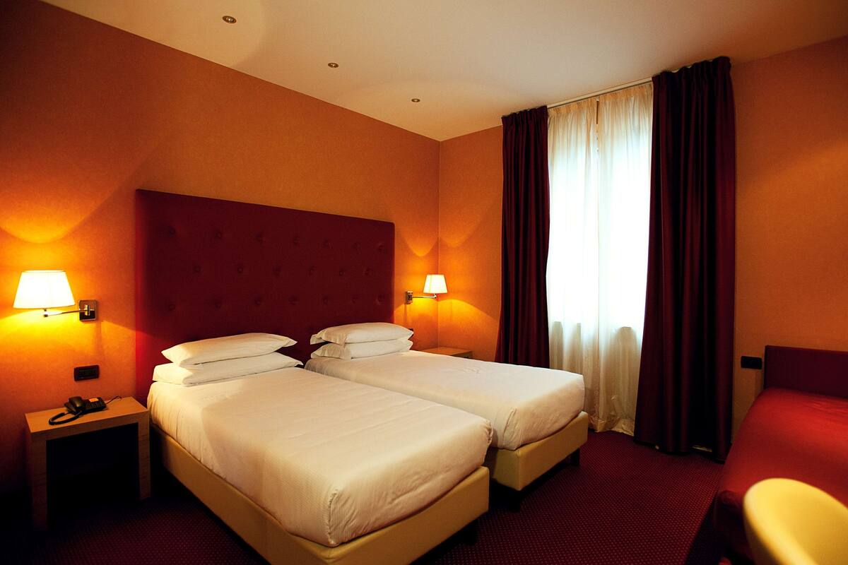 Habitación de hotel con dos camas, cabecera roja, dos lámparas de noche, mesita de noche con teléfono y una ventana con cortinas.
