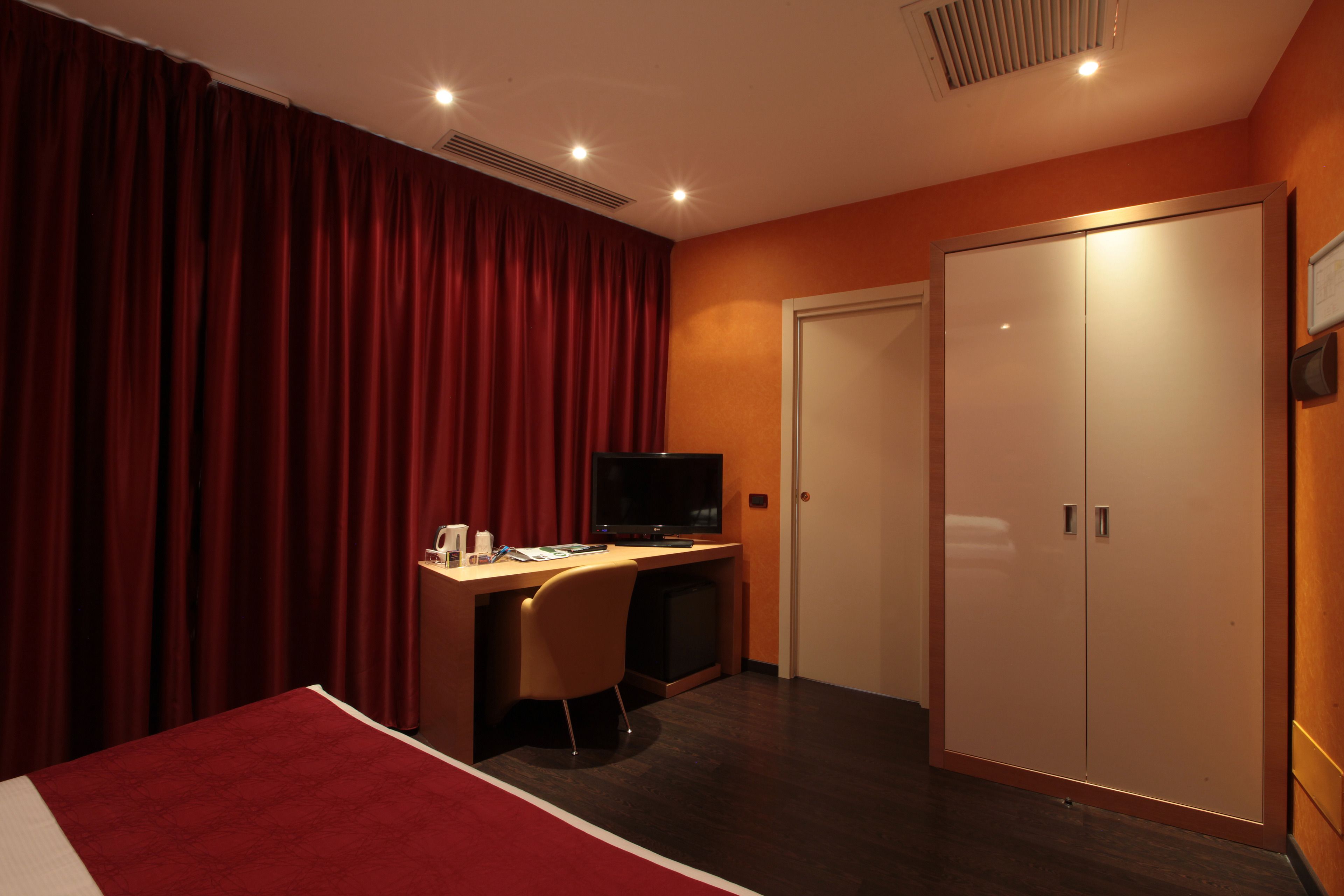 A hotel room with a bed, a desk with a television, a wardrobe, and red curtains.