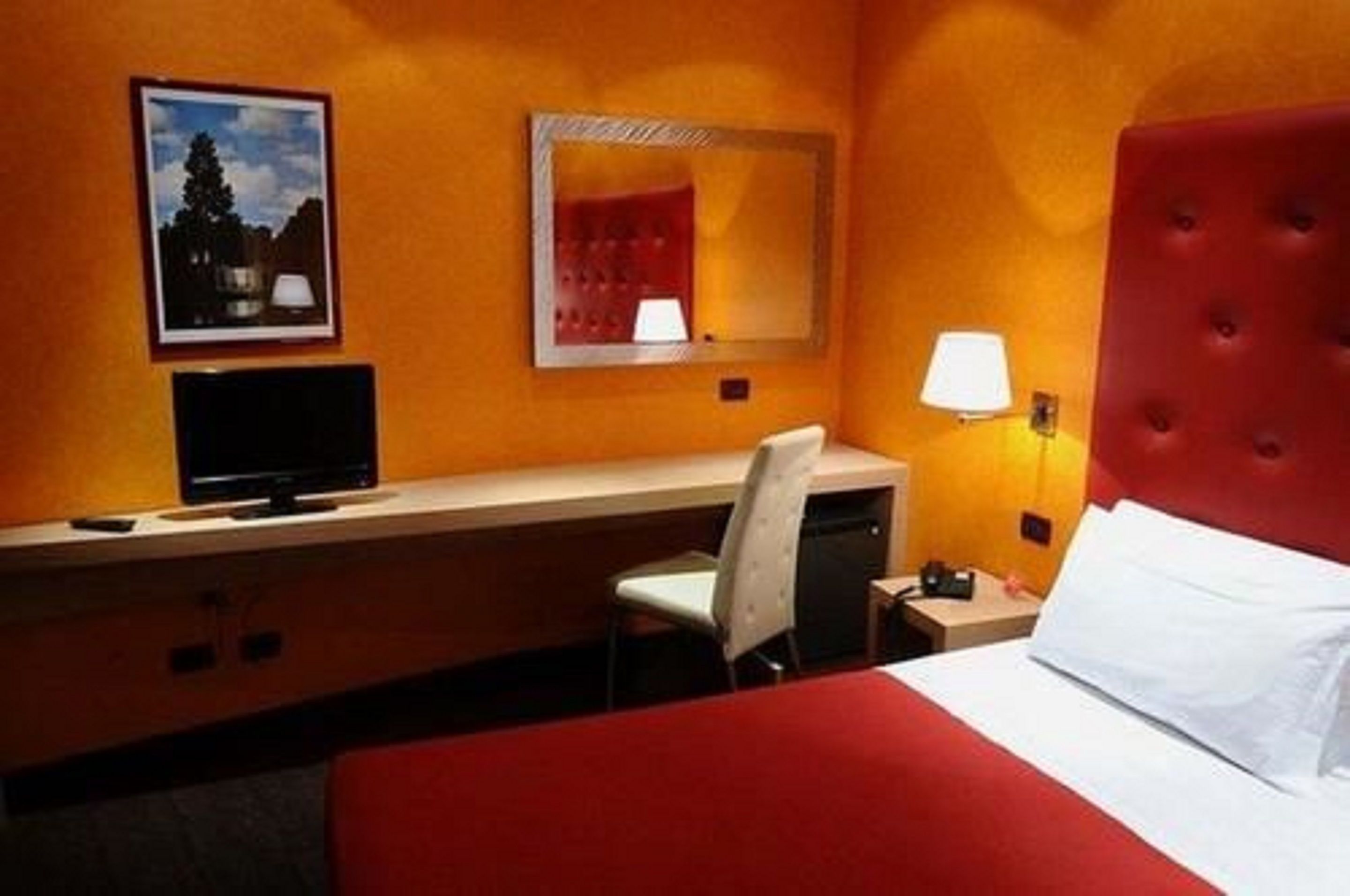 A hotel room with a bed, a desk with a chair, a television, a mirror, and a lamp.