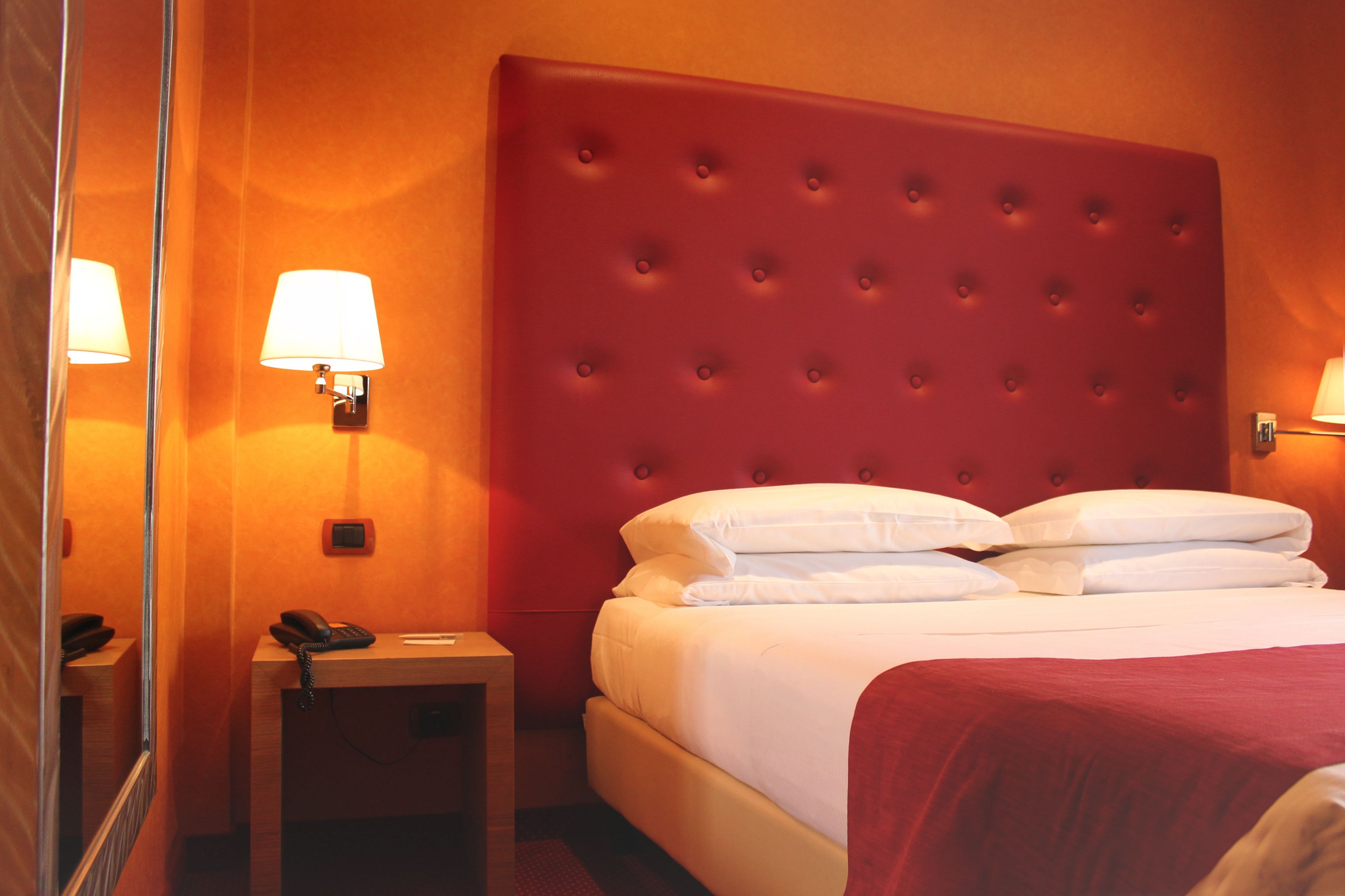Ein Hotelzimmer mit rotem, gepolstertem Kopfteil, weißer Bettwäsche, einer Nachttischlampe, einem Telefon und einem kleinen Tisch.