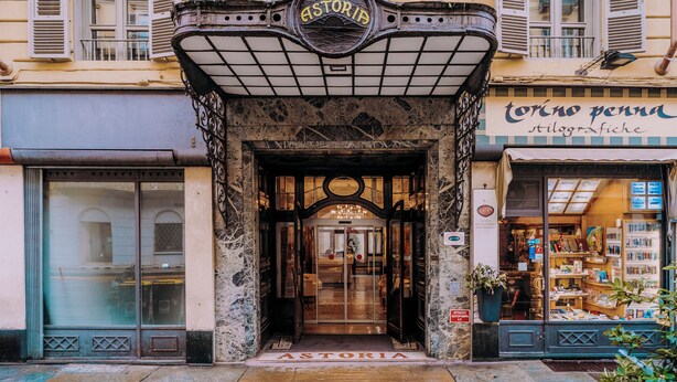 La entrada de un edificio con un letrero que dice "ASTORIA" y una tienda con un letrero que dice "torino penna stilografiche".