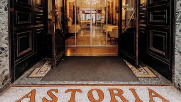 L'ingresso di un hotel con pavimento piastrellato che reca la scritta "ASTORIA" e una porta in vetro che dà sull'interno.
