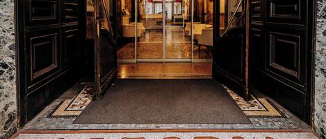 Entrada de hotel con suelo de baldosas que muestra el letrero "ASTORIA" y una puerta de vidrio que da al interior.