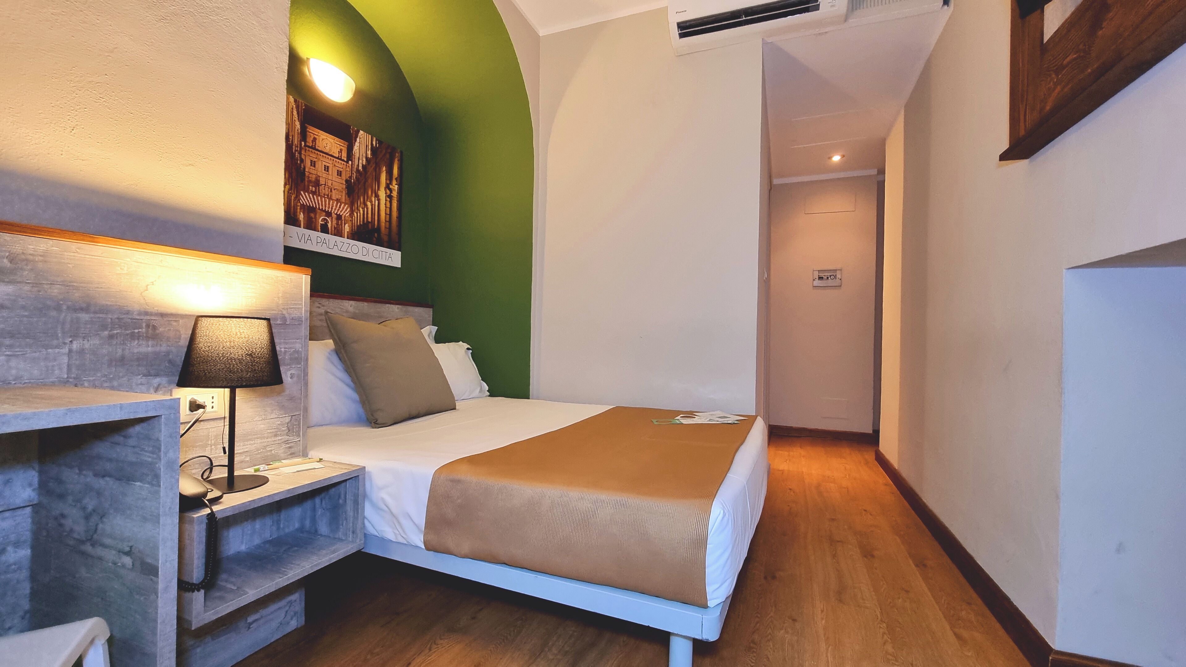 A small, narrow hotel room with a bed, bedside table, lamp, and a green accent wall.
