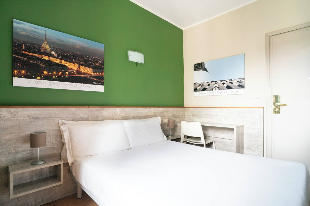 Habitación de hotel con cama, escritorio y dos sillas. Hay dos cuadros en la pared.