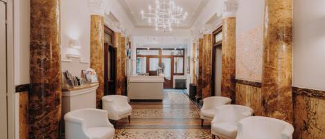 A hallway with marble columns, a chandelier, and white armchairs.