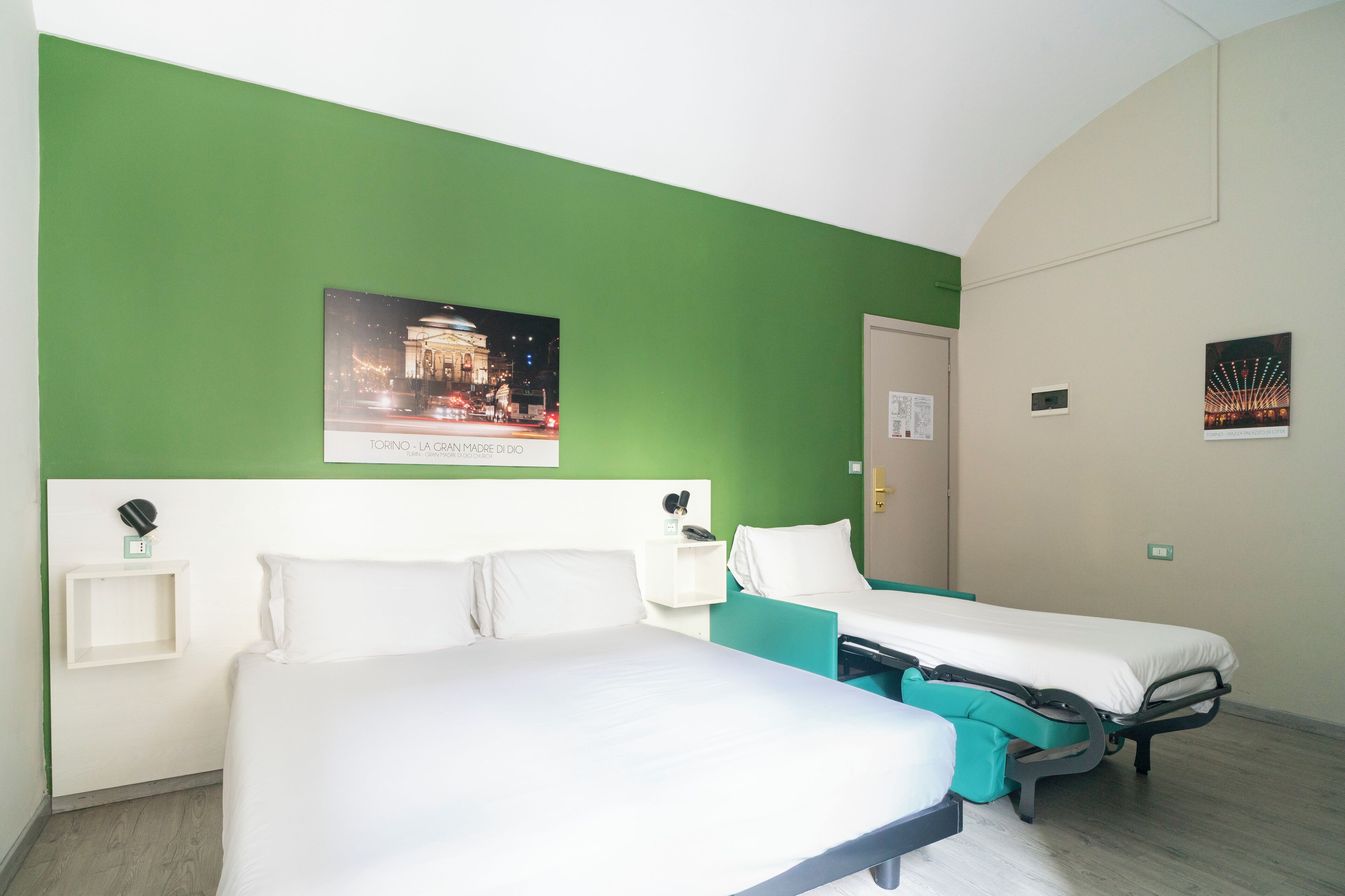 Une chambre d’hôtel avec deux lits simples, un mur d’accent de couleur verte, un tableau encadré représentant un paysage urbain et une petite table de chevet avec une lampe.