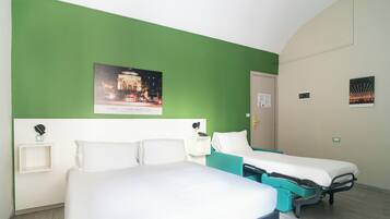 A hotel room with two single beds, a green accent wall, a framed picture of a cityscape, and a small bedside table with a lamp.