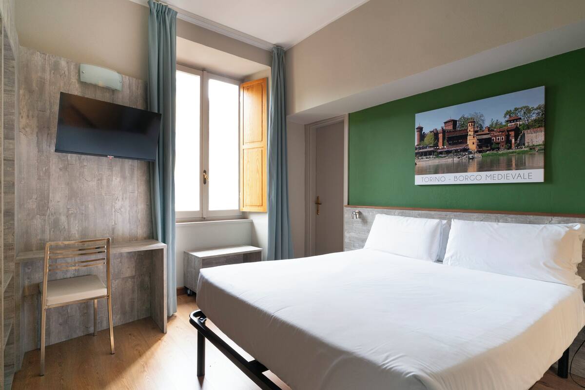 Un dormitorio con cama, escritorio y silla. Hay un cuadro en la pared y una ventana con cortinas.