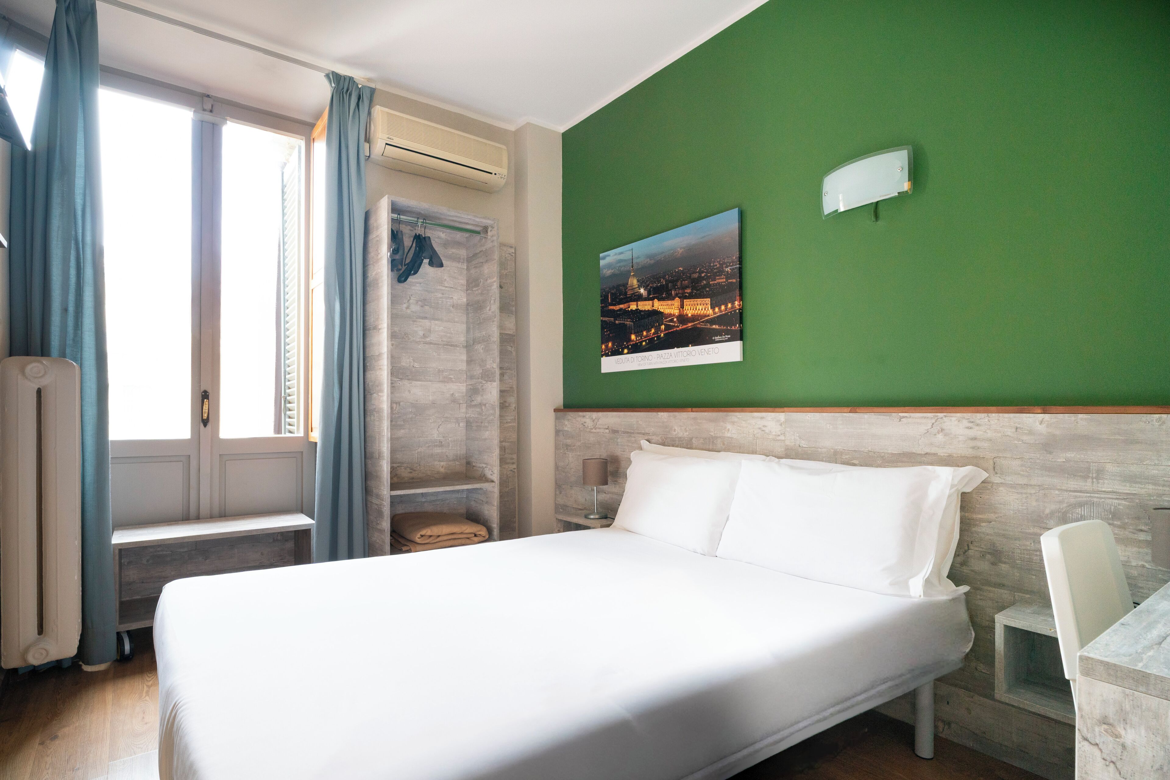 A hotel room with a bed, a desk, a chair, a window with curtains, a towel rack, and a picture on the wall.