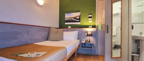 A hotel room with a bed, a nightstand, a lamp, a picture on the wall, and a bathroom visible through an open door.