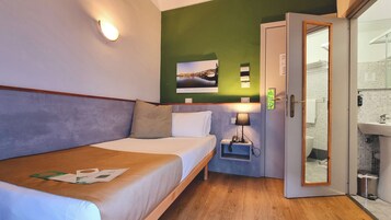 Habitación de hotel con cama, mesita de noche, lámpara, un cuadro en la pared y baño visible a través de una puerta abierta.