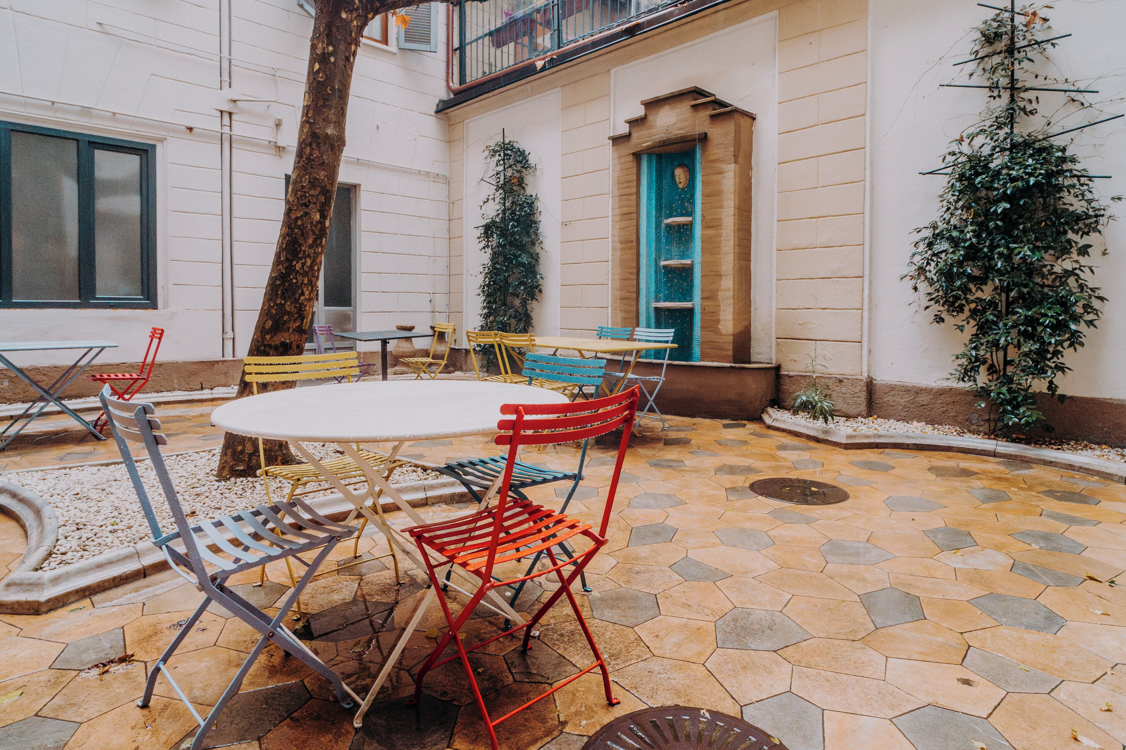 An outdoor patio area with a round table and chairs, a tree, and a building with a blue door and windows.