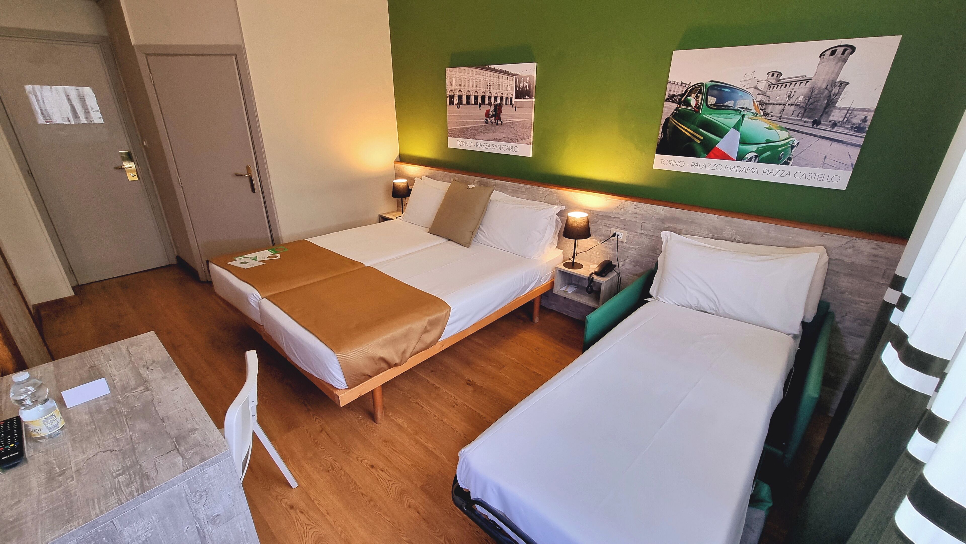 Camera d'albergo con due letti, un tavolino e una sedia. Sono presenti lampade a parete e un quadro raffigurante un edificio.