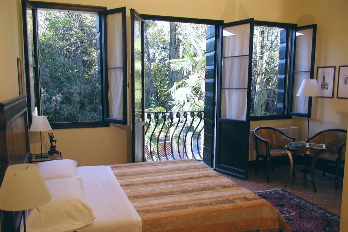 A hotel room with a bed, a desk, a chair, and a view of the outdoors through open windows.