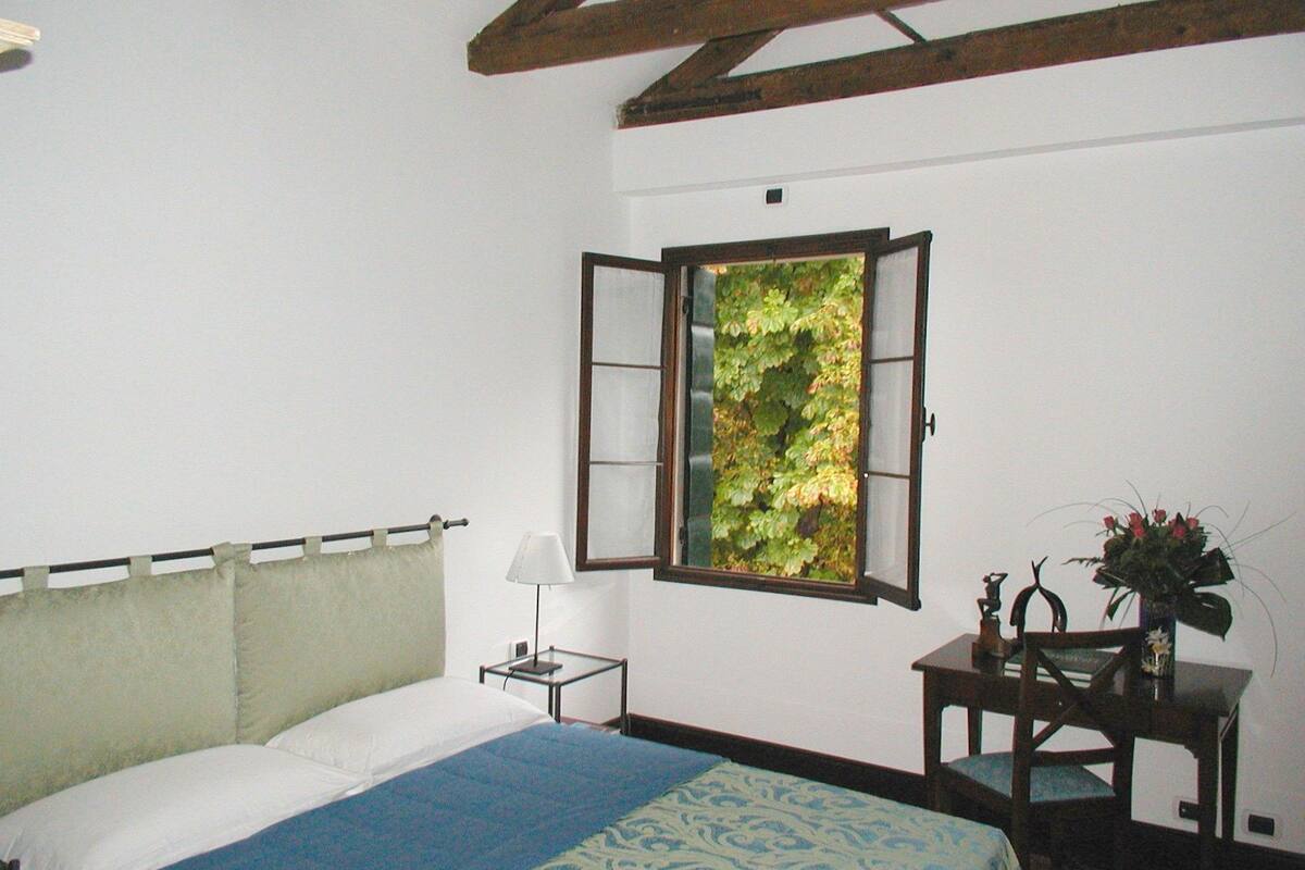 A bedroom with a bed, a nightstand, a desk, and a window with a view of greenery.