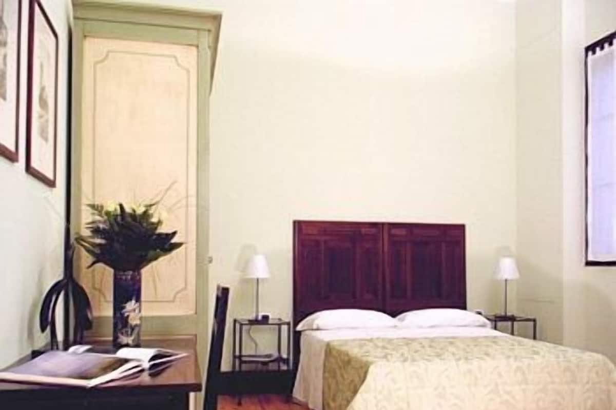A bedroom with a wooden bed, two bedside tables with lamps, a chair, a desk with a book, and a potted plant.