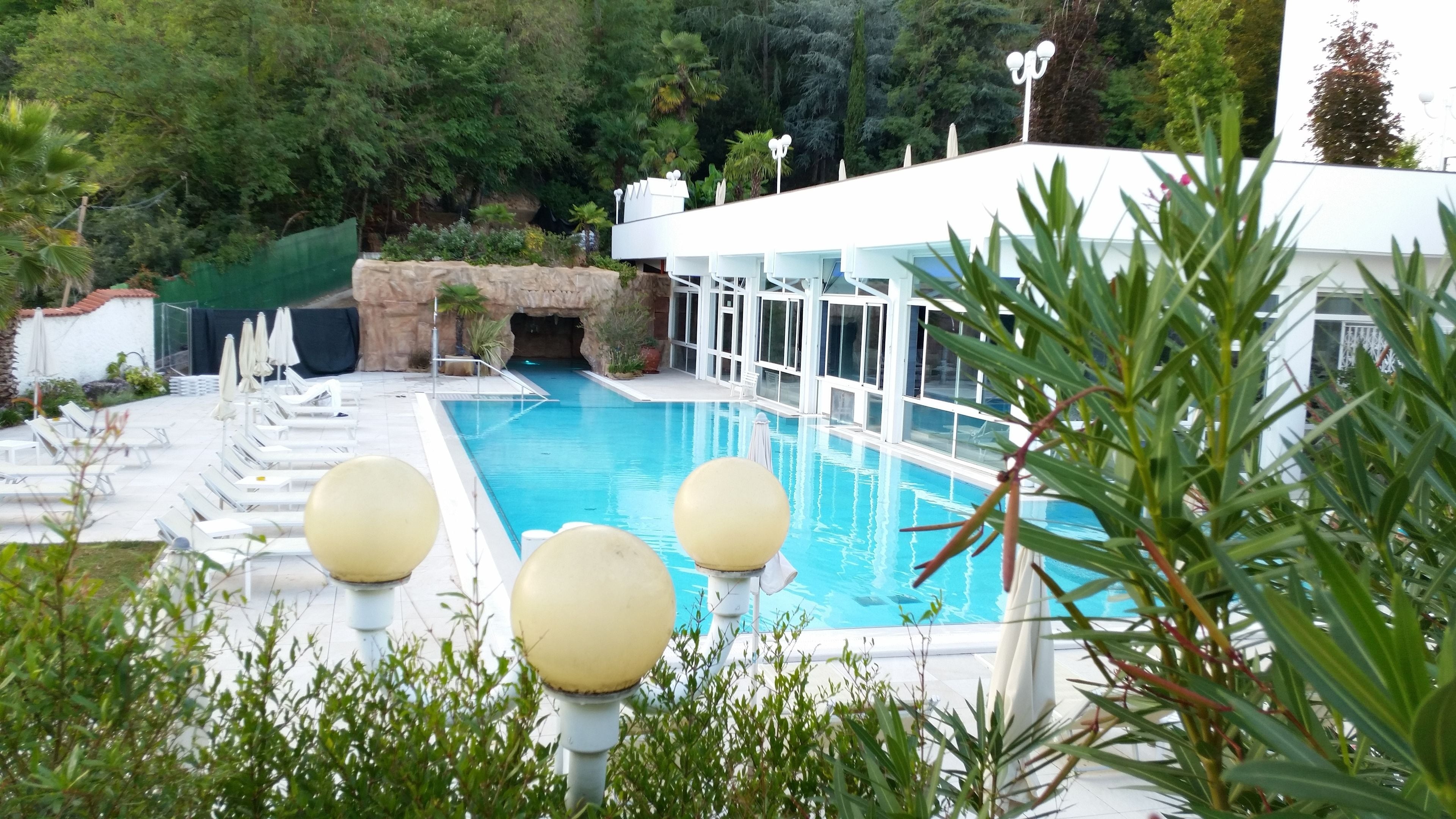 A swimming pool with lounge chairs and umbrellas, surrounded by greenery and a building with large windows.