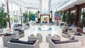 A spacious indoor area with wicker furniture arranged around a central pool, large windows, and indoor plants. - Hotel Terme Millepini (Montegrotto Terme)