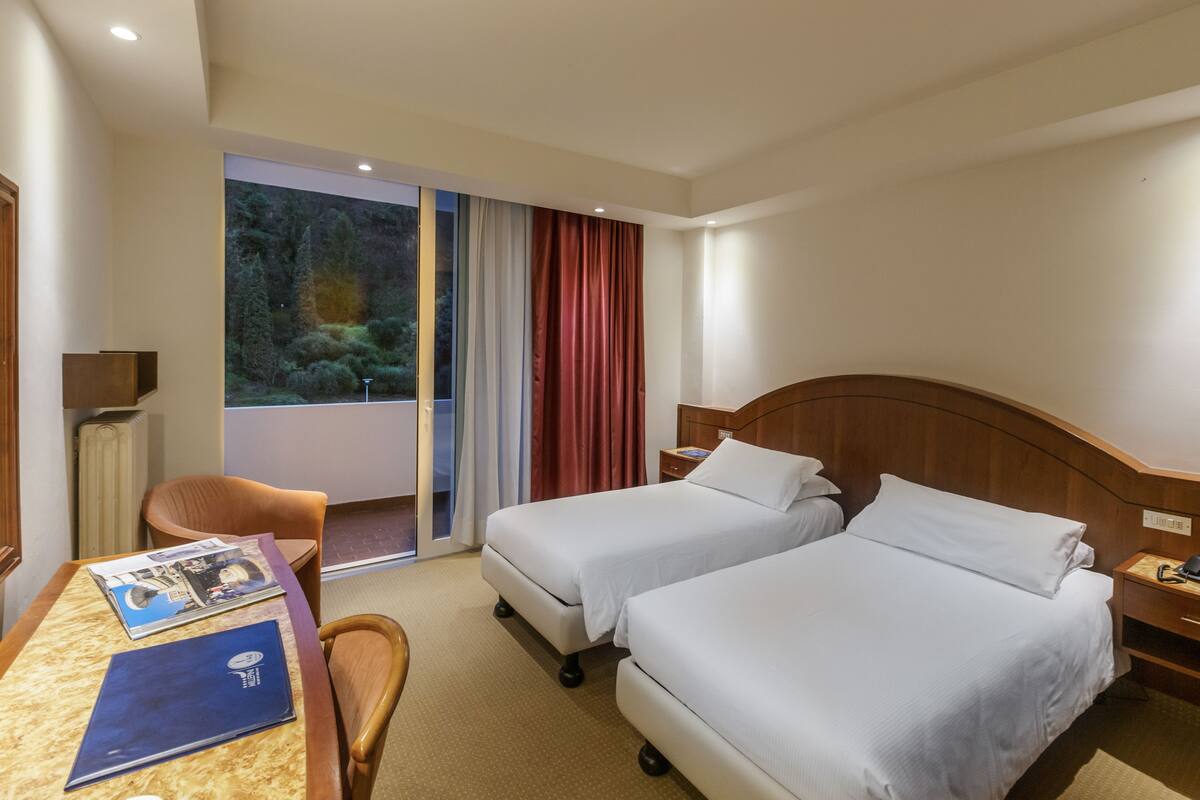 A hotel room with two beds, a desk with a chair, a window with curtains, and a view of greenery outside.