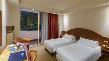 A hotel room with two beds, a desk with a chair, a window with curtains, and a view of greenery outside.