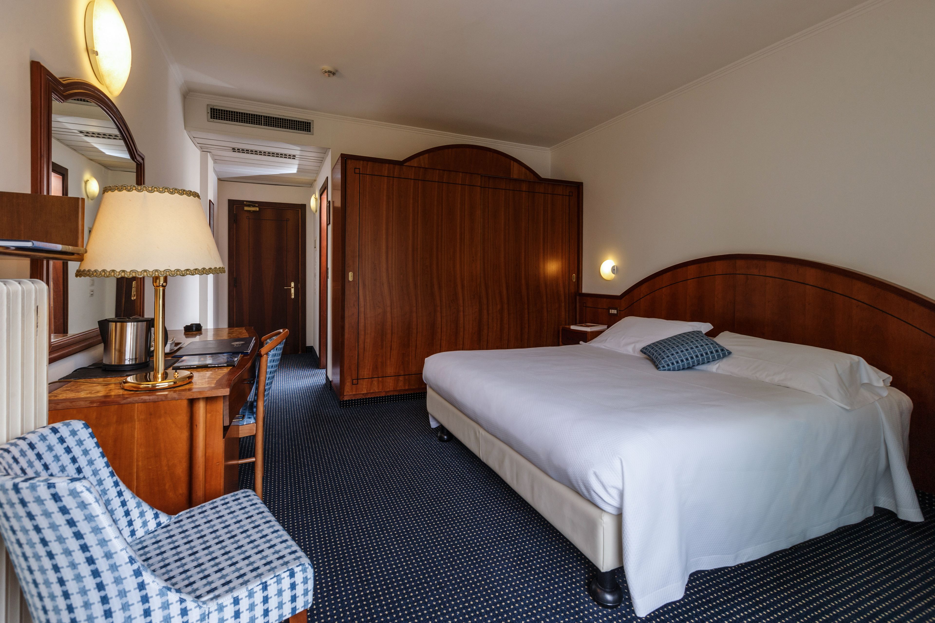 A hotel room with a bed, a desk with a lamp, a chair, a wardrobe, and a mirror.