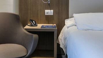 Una camera d'albergo moderna con una parete in legno decorativa, un comodino con telefono e libro, una sedia grigia e un letto con lenzuola bianche.