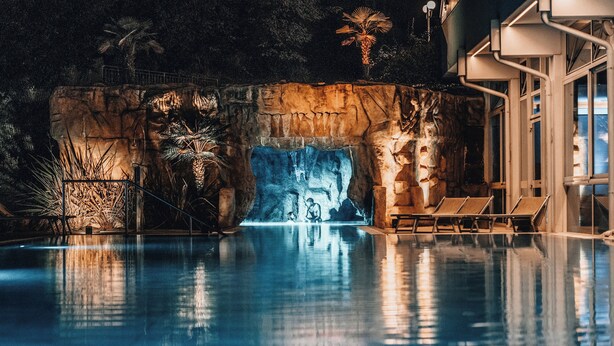 A swimming pool with a rock wall, palm trees, and lounge chairs. - Hotel Terme Millepini (Montegrotto Terme)