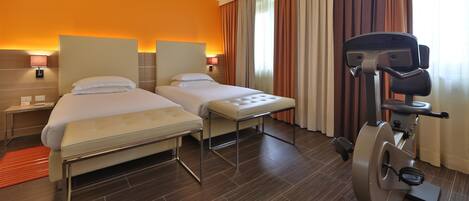Habitación de hotel con dos camas, una cinta de correr y una mesita de noche con lámpara.