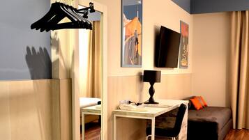 Een hotelkamer met een houten vloer, een bureau met een lamp, een bed met een oranje kussen, een televisie aan de muur en een kapstok met kleding.