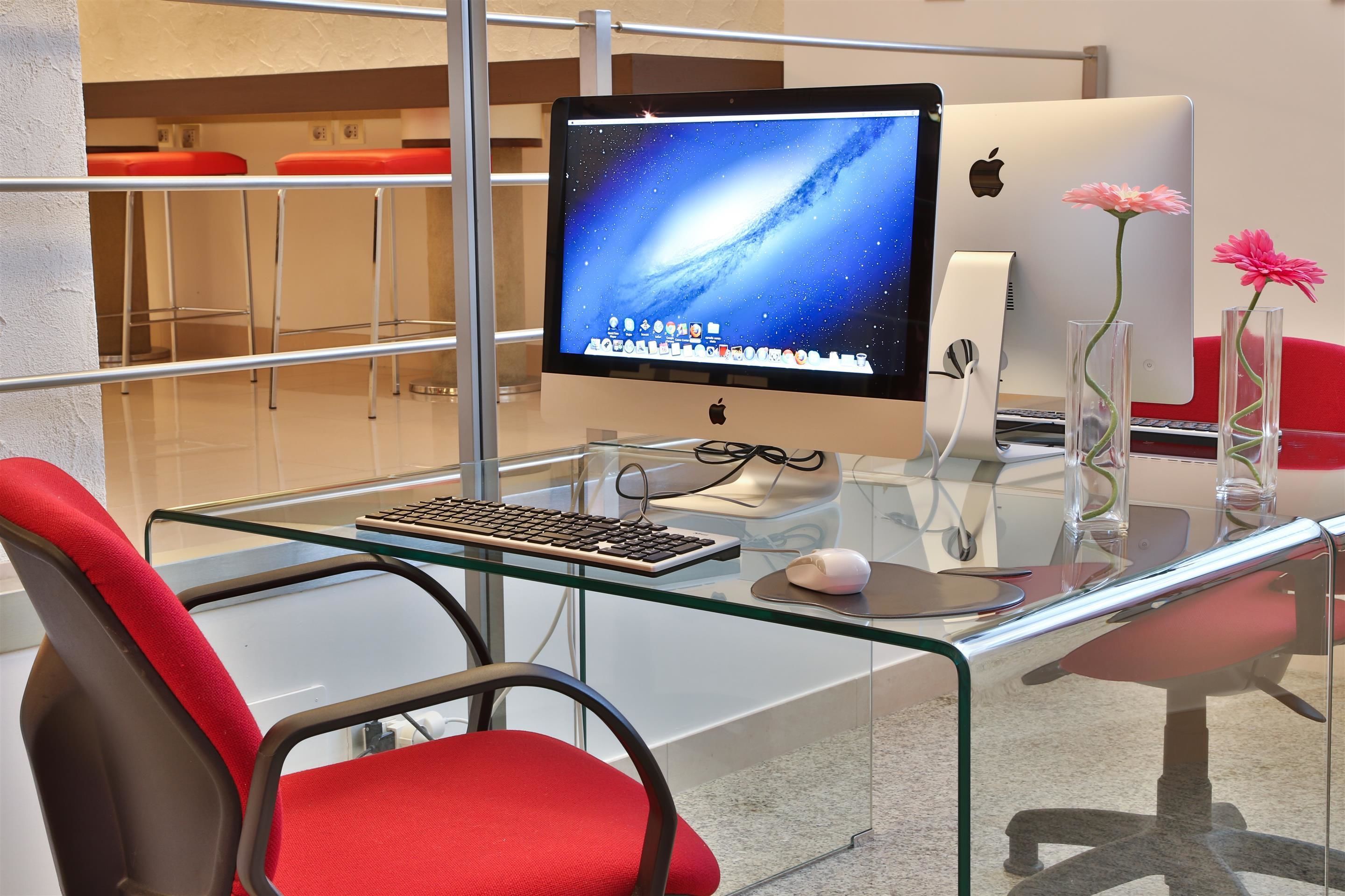 A modern office desk with a computer, keyboard, and mouse. There are two red chairs and a glass table.