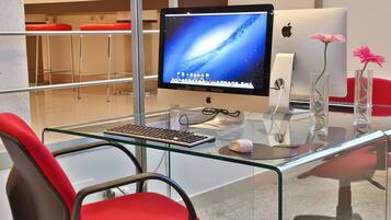 A modern office desk with a computer, keyboard, and mouse. There are two red chairs and a glass table.