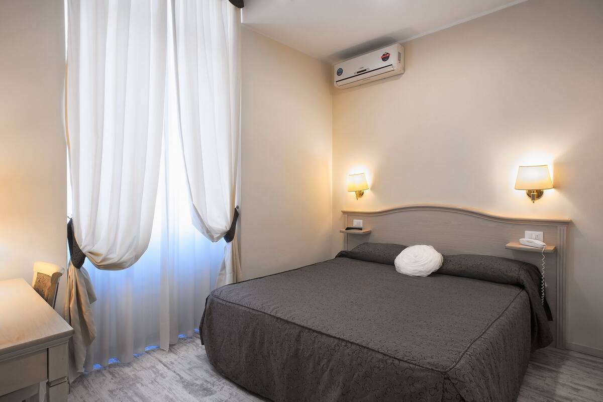 A hotel room with a bed, bedside lamps, an air conditioning unit, and a desk with a lamp.