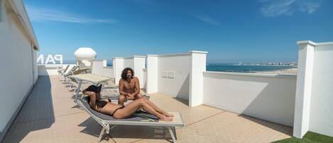 A rooftop lounge area with sun loungers, a pool, and a view of the sea.