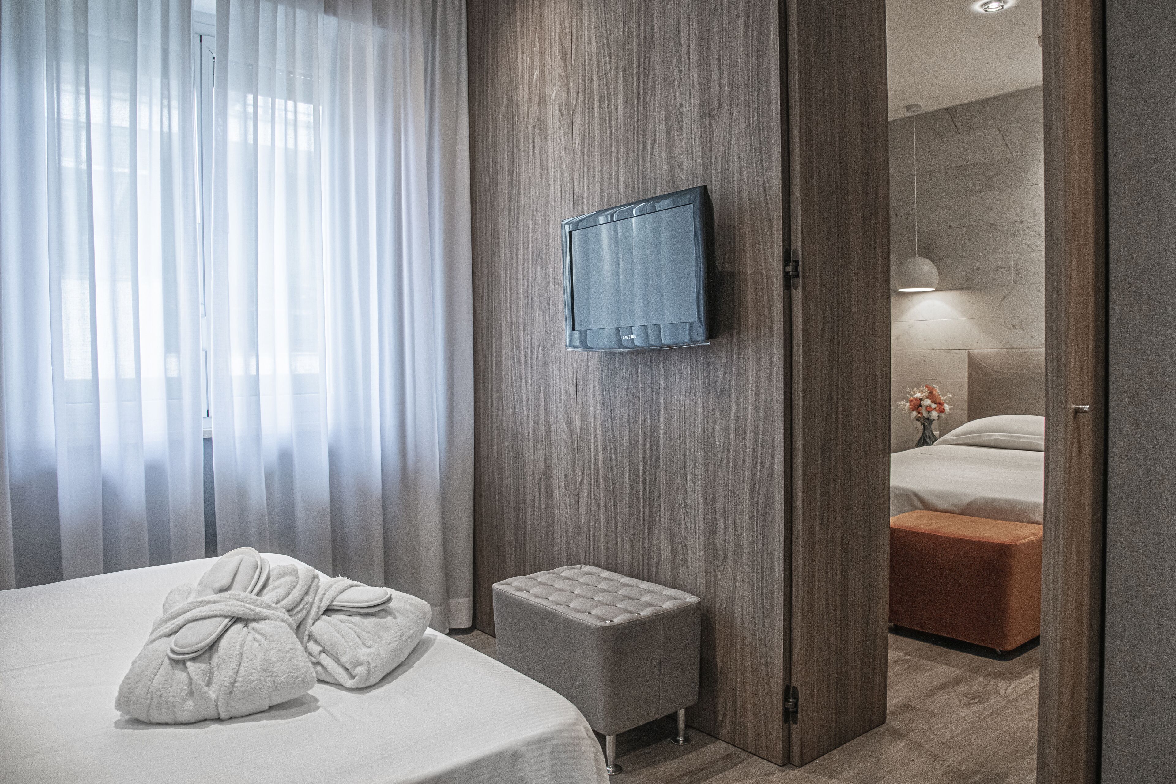 A hotel room with a bed, a TV mounted on the wall, a small ottoman, and a bathroom visible through an open door.