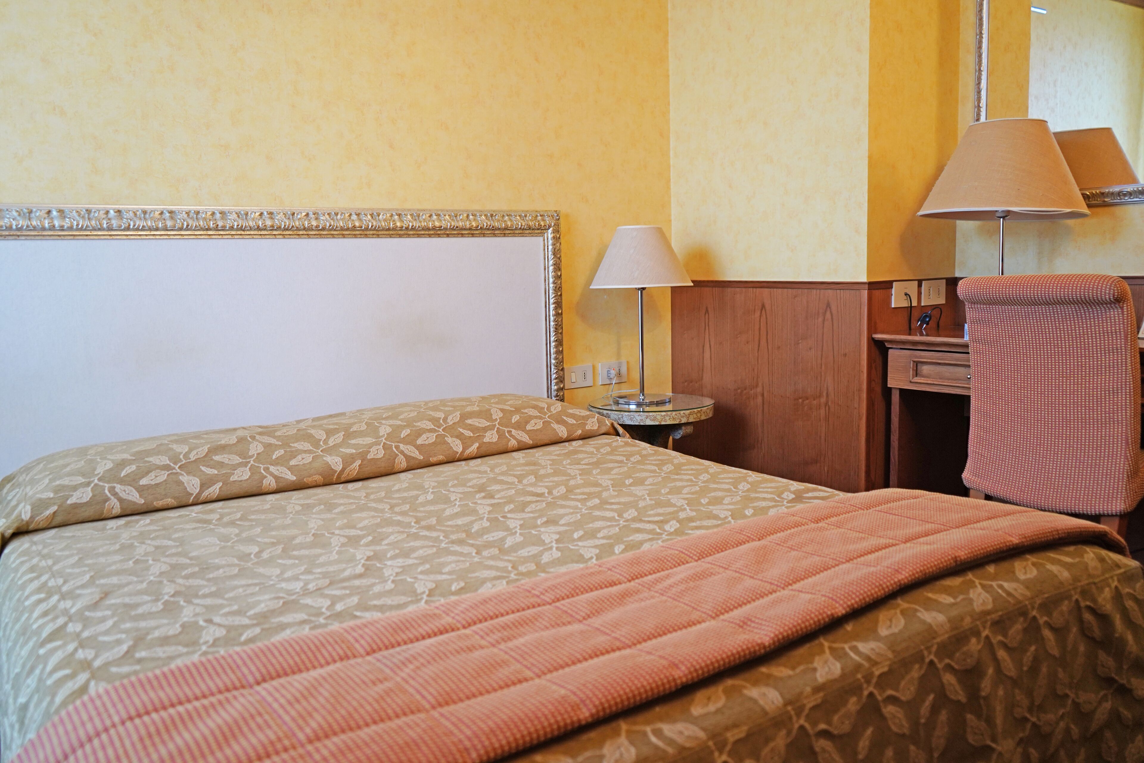 Quarto de hotel com cama, escrivaninha, luminária e cadeira.