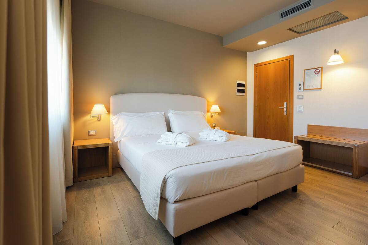 A neatly made bed with white linens and pillows, two bedside lamps, a wooden bench, and a wooden door.