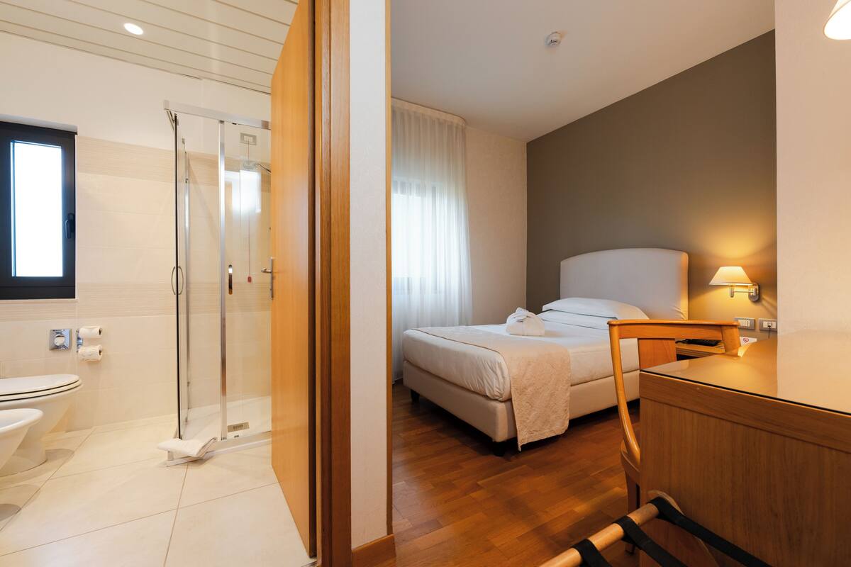 A hotel room with a shower, toilet, bed, bedside table, and a lamp.