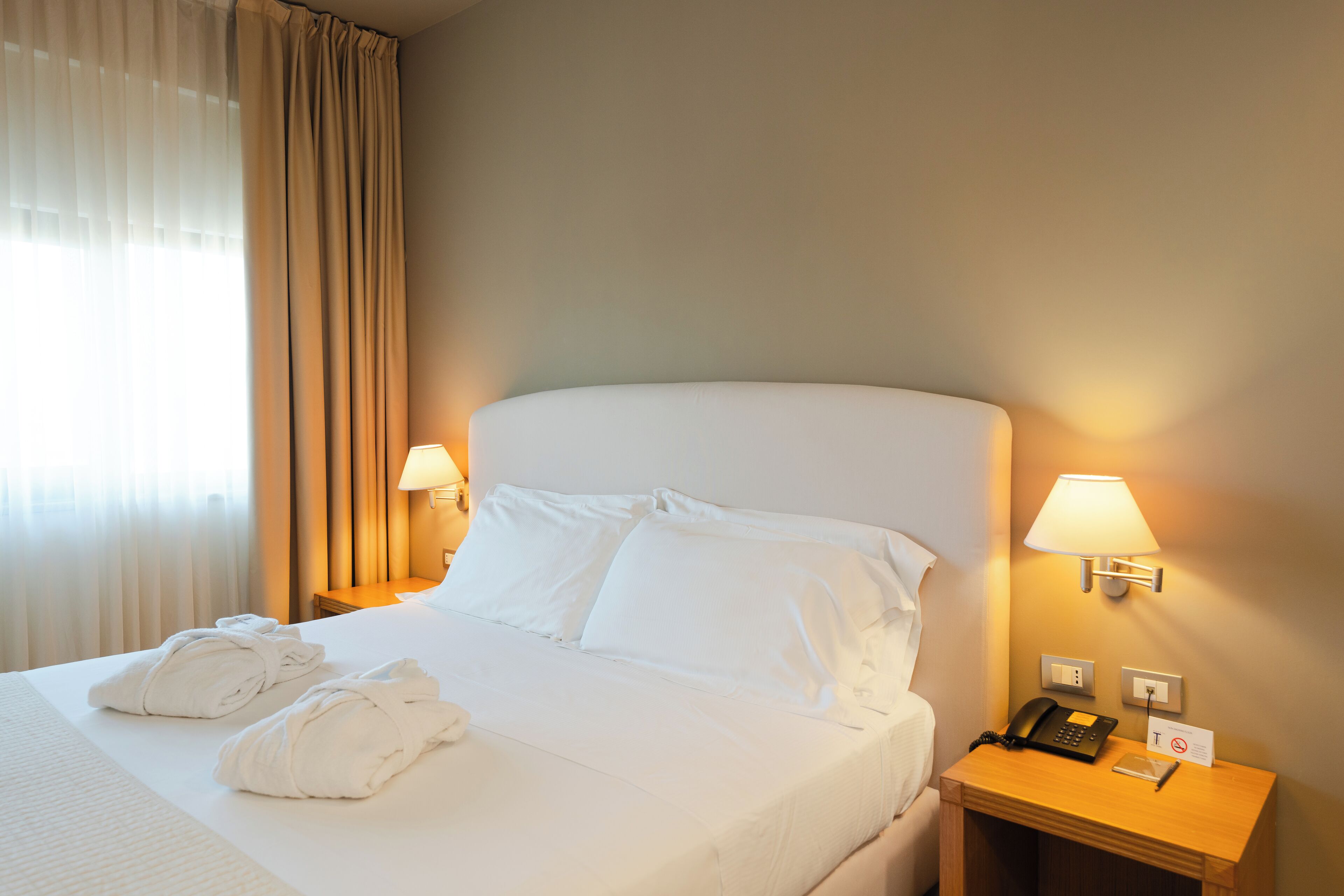 A hotel room with a neatly made bed, two bedside lamps, a bedside phone, and a sign indicating no smoking.