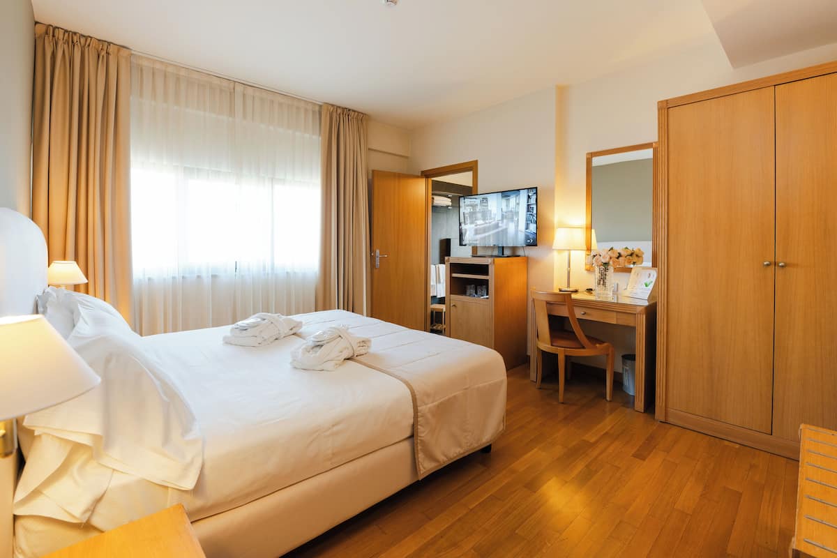 A hotel room with a large bed, a desk with a chair, a television, and a wardrobe.