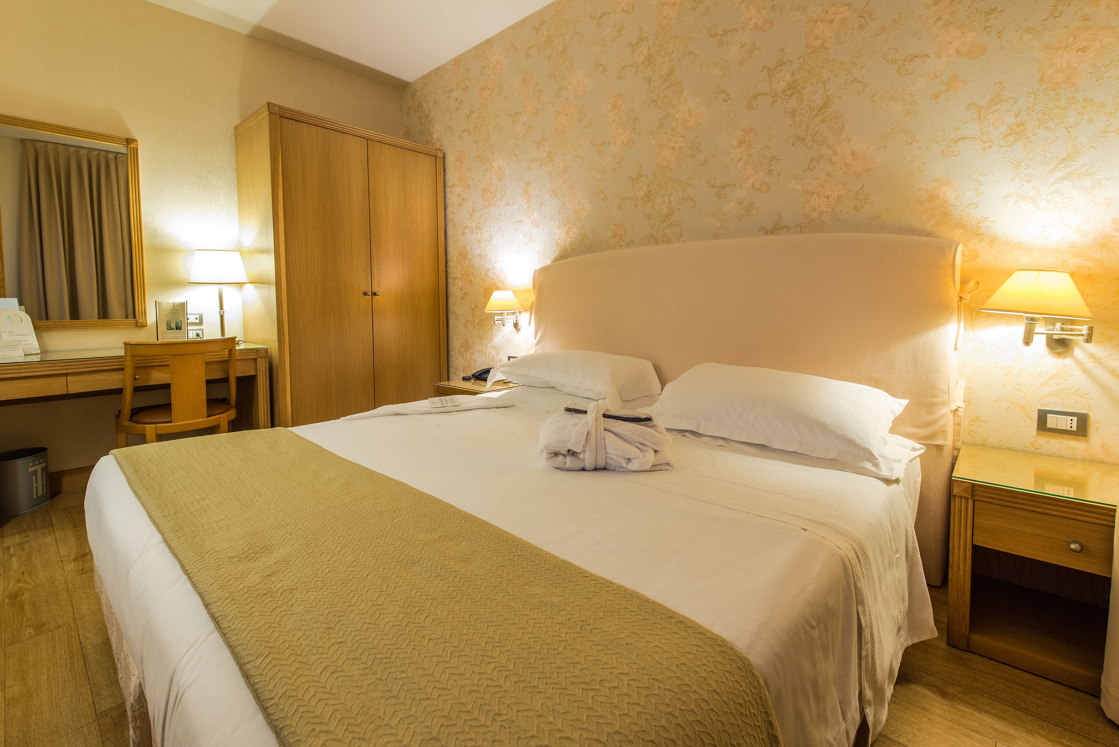 A hotel room with a large bed, two bedside lamps, a desk with a chair, a wardrobe, and a mirror.