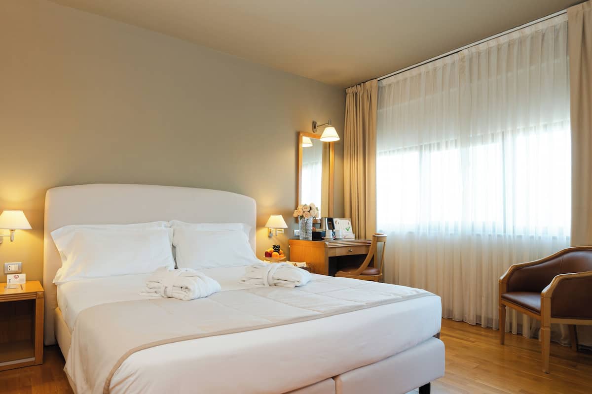 A hotel room with a large bed, a desk with a chair, a mirror, and a window with curtains.