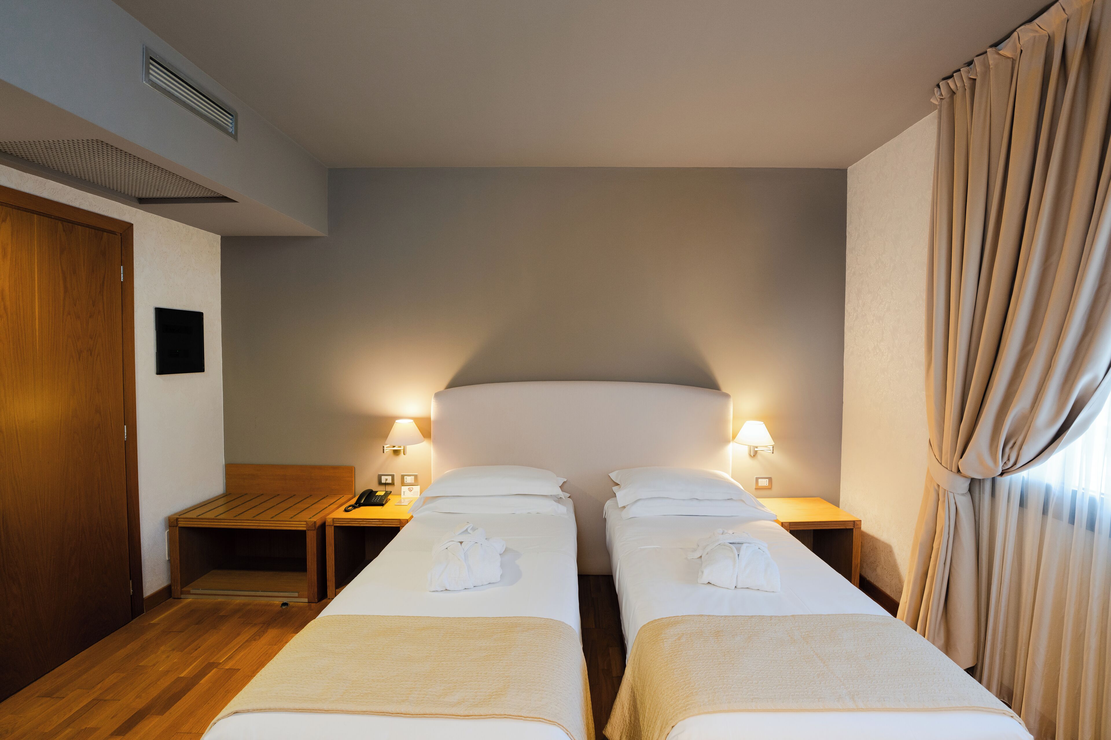 A hotel room with two single beds, wooden headboards, bedside tables with lamps, and a wooden floor.