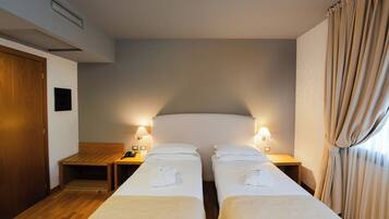A hotel room with two single beds, wooden headboards, bedside tables with lamps, and a wooden floor.