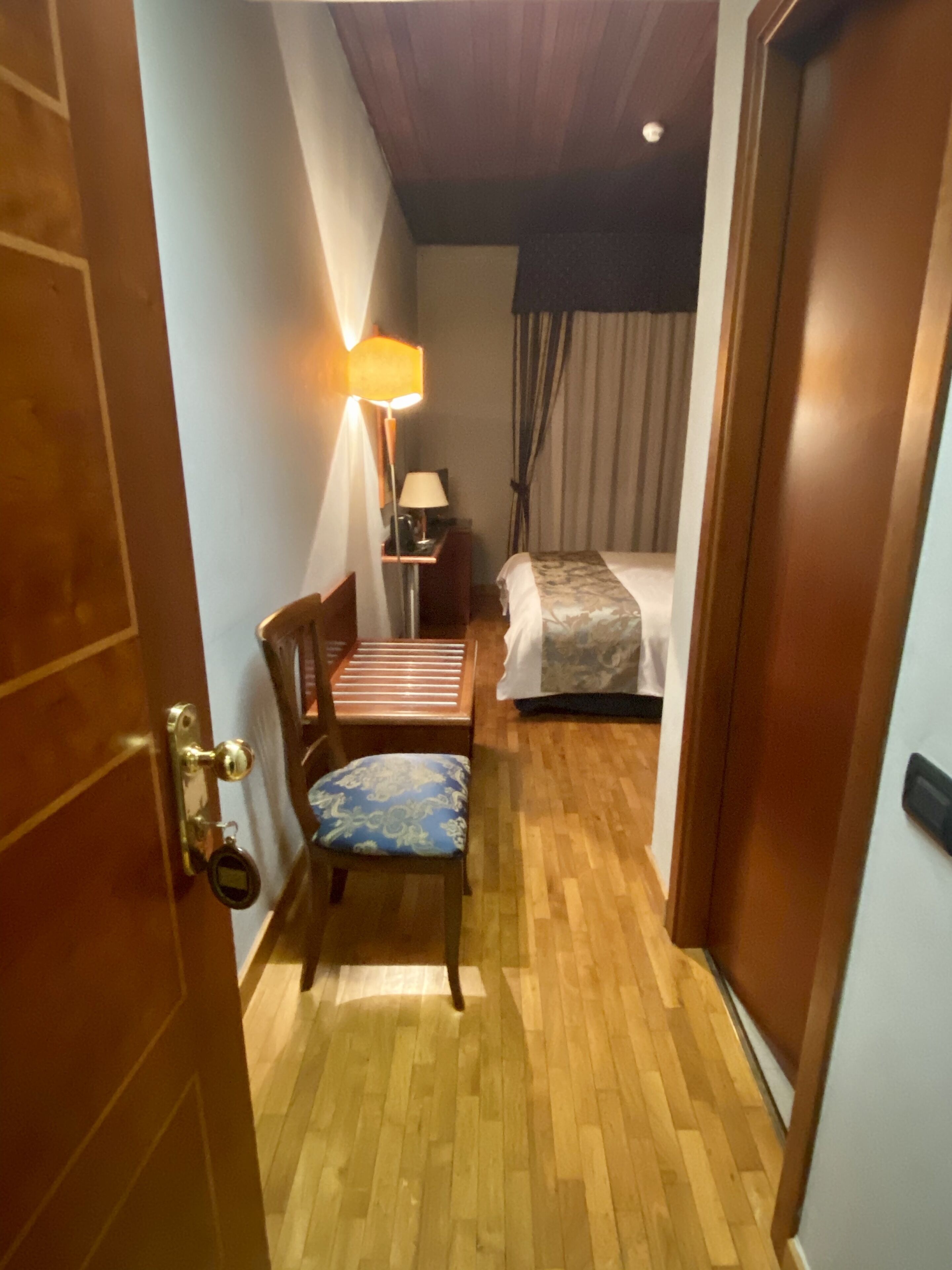 Habitación de hotel con piso de madera, cama con colcha estampada, una silla con cojín, mesita de noche con lámpara y puerta con picaporte de latón.