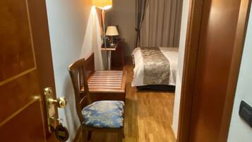 A hotel room with a wooden floor, a bed with a patterned bedspread, a chair with a cushion, a nightstand with a lamp, and a door with a brass handle.