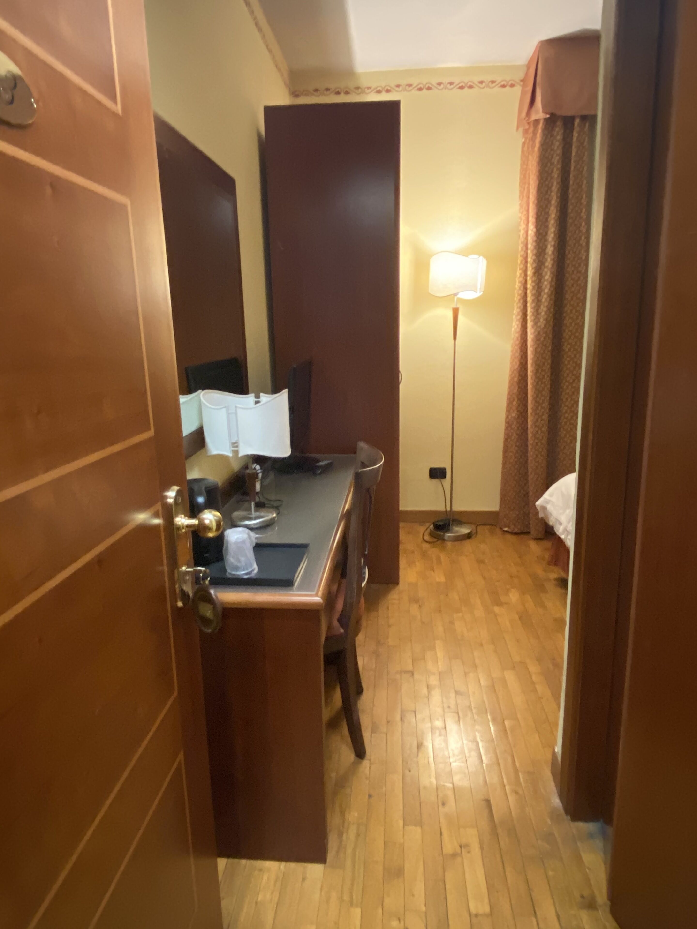 A hotel room with a wooden floor, a desk with a chair, a bed, a nightstand, a lamp, and a window with curtains.