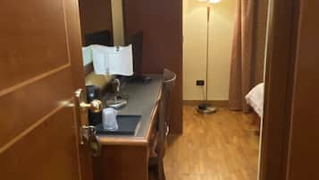 A hotel room with a wooden floor, a desk with a chair, a bed, a nightstand, a lamp, and a window with curtains.