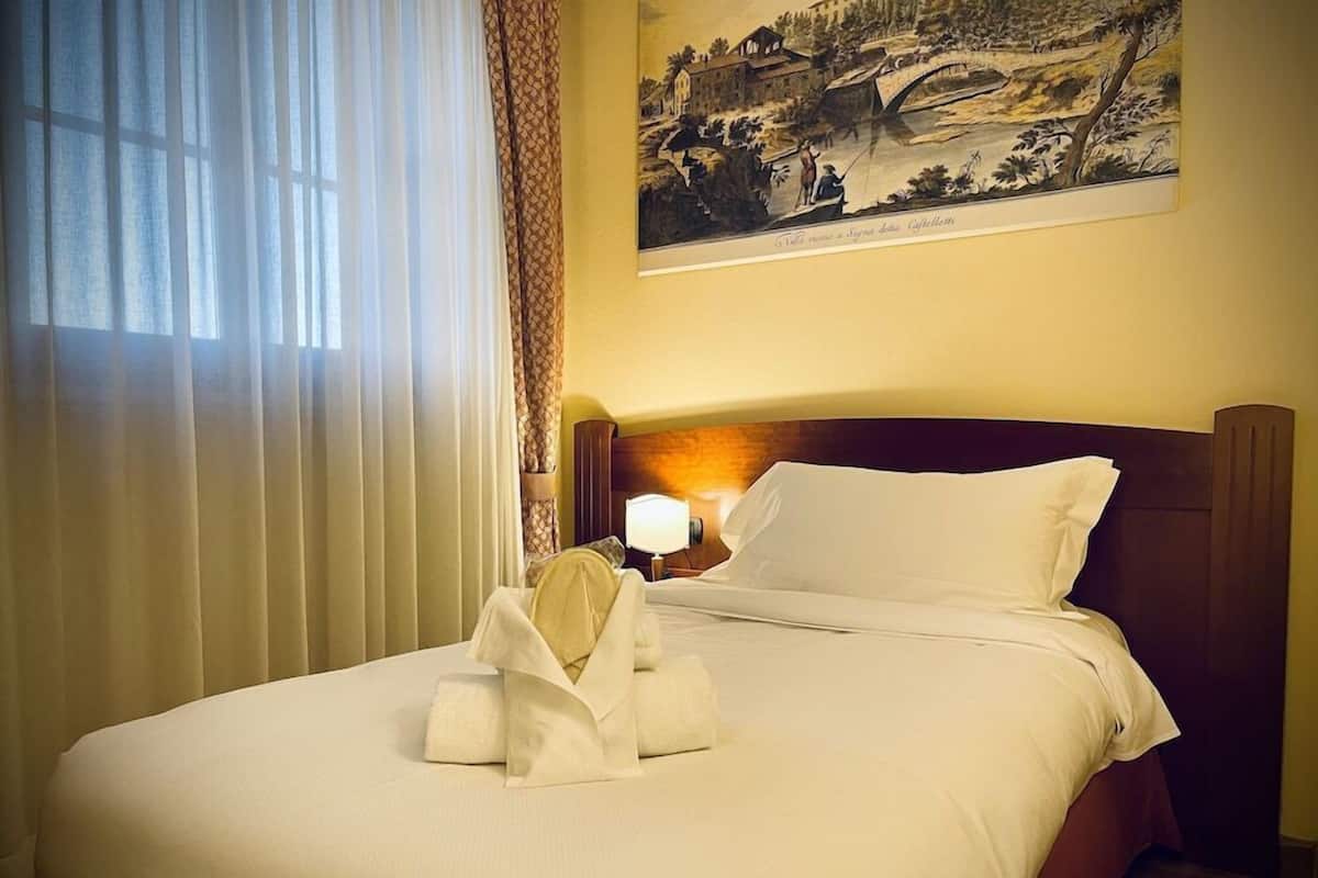 A hotel room with a neatly made bed, a lamp on the nightstand, and a painting on the wall.